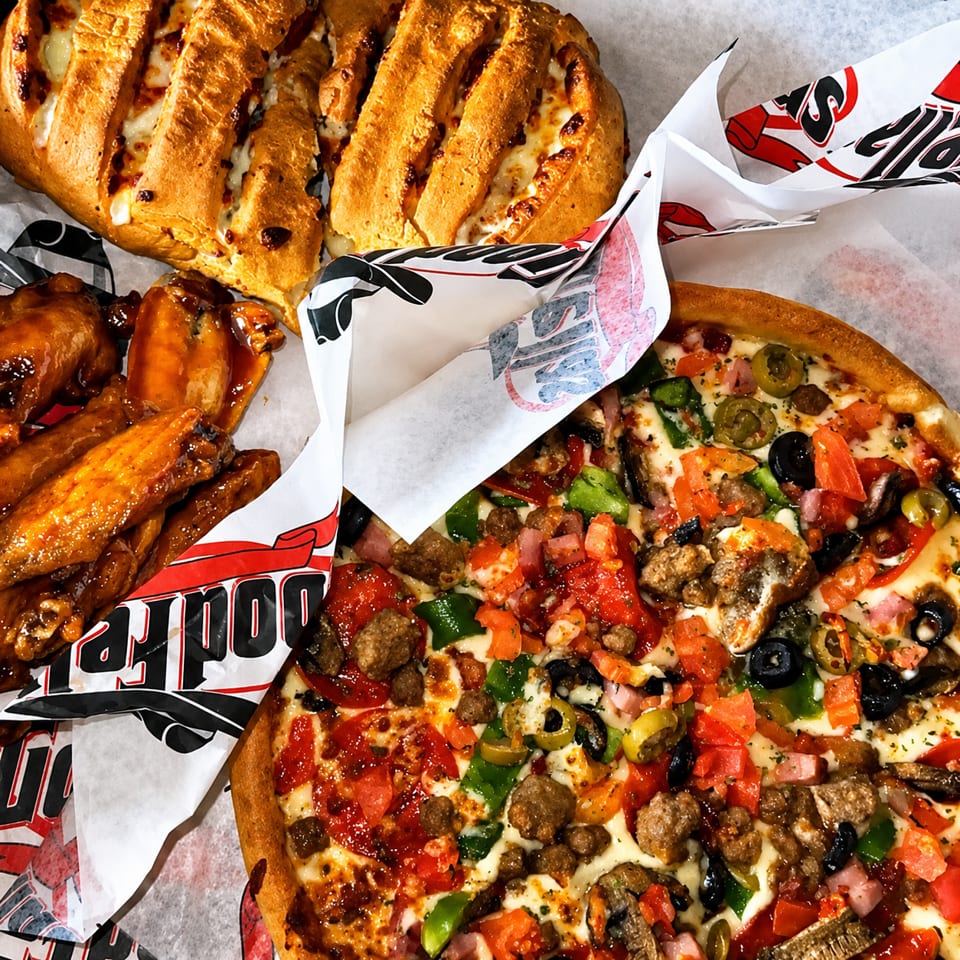 Loaded meat-topped pizza, cheesy breadsticks, and saucy chicken wings arranged on branded restaurant paper.